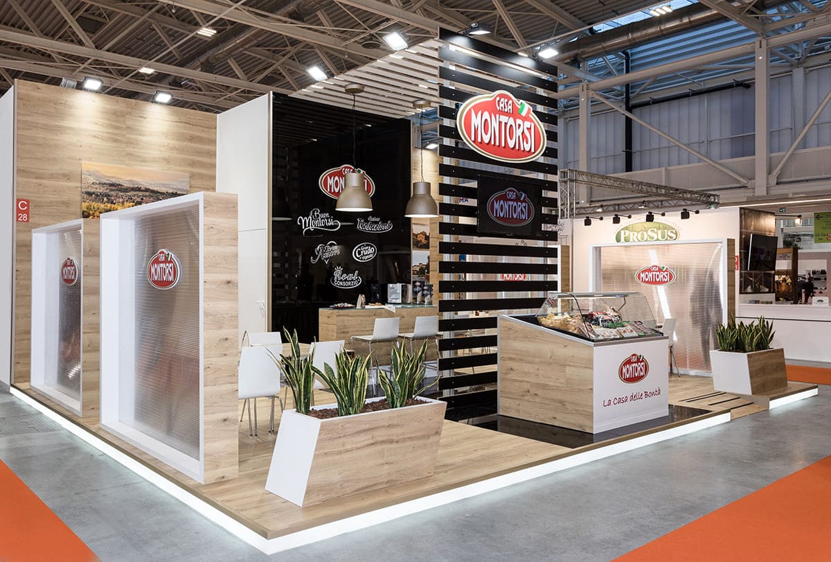 Allestimenti fieristici Cibus Parma casi reali: l'allestimento stand realizzato per Casa Montorsi L'immagine mostra un caso reale: l'allestimento dello stand per fiera Cibus di Parma realizzato per Casa Montorsi SpA visto dall'esterno. Si tratta di uno stand per fiere allestito su un pavimento rialzato, con zoccolo retroilluminato, rivestito in rovere naturale. La struttura è formata da una parete di fondo rivestita anch'essa in rovere naturale e con un box magazzino centrale rifinito in bianco e nero lucido. La zona centrale è caratterizzata dalla presenza di una struttura a ponte, lavorata a doghe di colore bianco e nero. Ai lati di questa struttura si trovano le due zone trattative con tavoli che sono schermati da diaframmi semitrasparenti. Completano l'allestimento di questo stand un'esposizione di prodotti in vetrina refrigerata frontale ed un paio di separé con piante verdi.