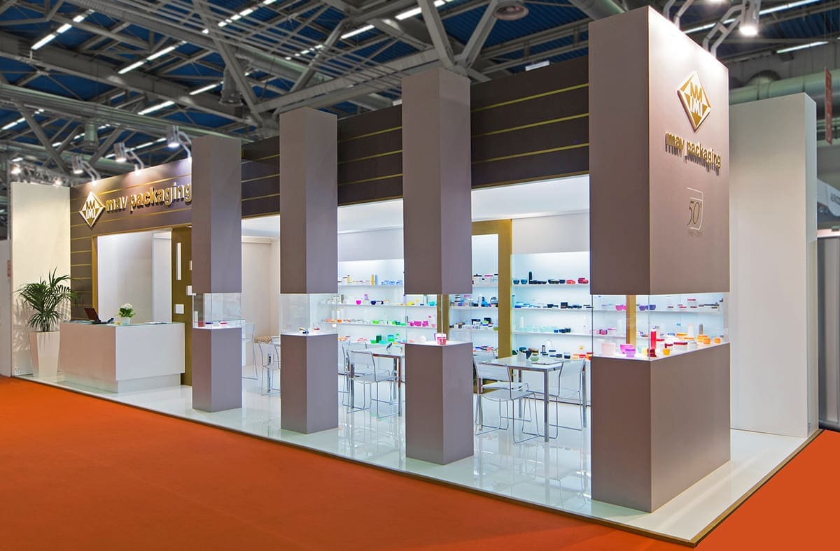 L'immagine mostra un caso reale: l'allestimento dello stand per fiere Cosmoprof Cosmopack di Bologna realizzato per MAV Packaging visto dall'esterno. Si tratta di uno stand per fiere allestito su un pavimento rialzato laccato bianco lucido. La struttura è formata da una parete di fondo bianca con nicchie espositive retroilluminate; poi una zona riservata sulla sinistra con box ripostiglio e bar; infine una struttura frontale fatta di vetrine a colonna collegate tra loro per mezzo di un frontone trasversale. All'interno della spazio ricreato si notano la zona tavoli per incontri e la zona del bar con il banco somministrazione.