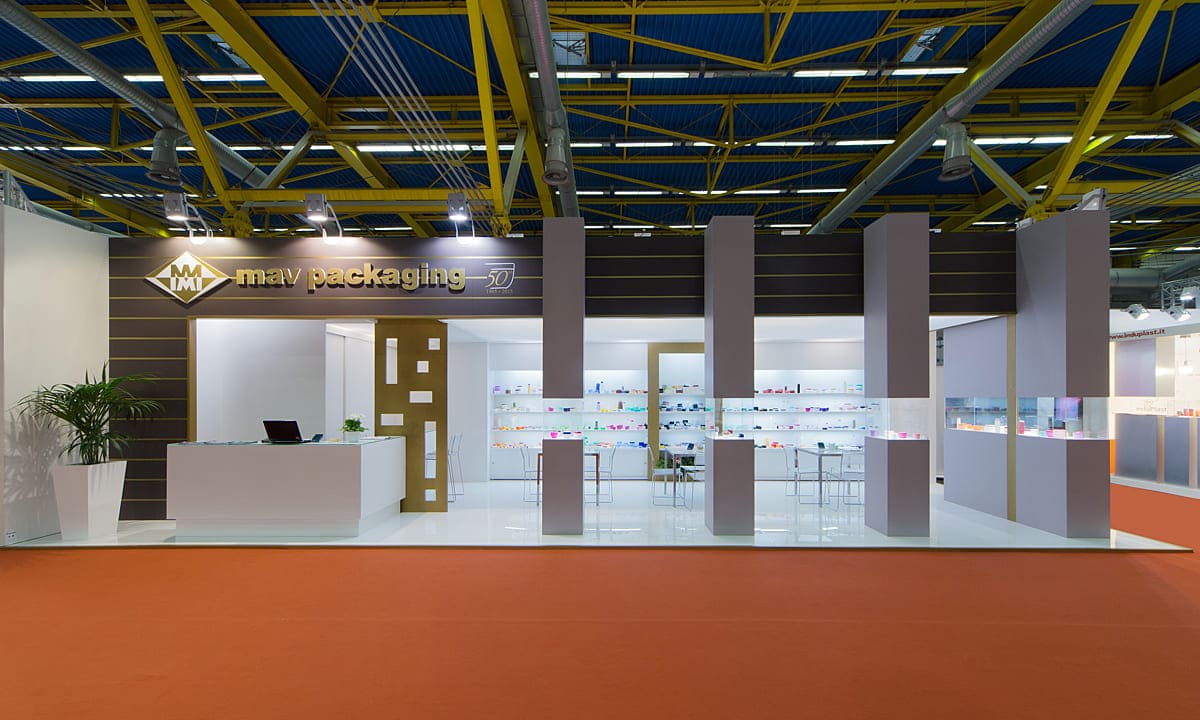 L'immagine mostra un caso reale: l'allestimento dello stand per fiere Cosmoprof Cosmopack di Bologna realizzato per MAV Packaging visto dall'esterno. Si tratta di uno stand per fiere allestito su un pavimento rialzato laccato bianco lucido. La struttura è formata da una parete di fondo bianca con nicchie espositive retroilluminate; poi una zona riservata sulla sinistra con box ripostiglio e bar; infine una struttura frontale fatta di vetrine a colonna collegate tra loro per mezzo di un frontone trasversale. All'interno della spazio ricreato si notano la zona tavoli per incontri e la zona del bar con il banco somministrazione.