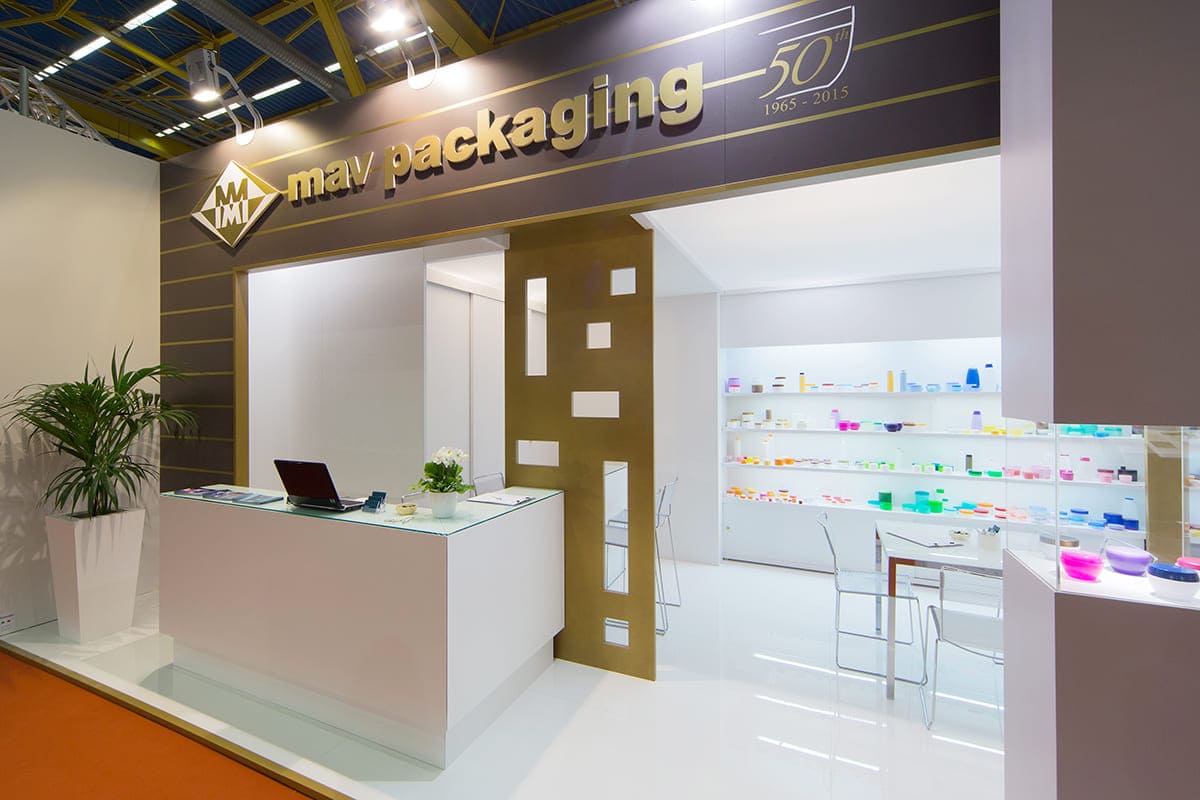 L'immagine mostra un caso reale: l'allestimento dello stand per fiere Cosmoprof Cosmopack di Bologna realizzato per MAV Packaging visto dall'esterno. Si tratta di uno stand per fiere allestito su un pavimento rialzato laccato bianco lucido. La struttura è formata da una parete di fondo bianca con nicchie espositive retroilluminate; poi una zona riservata sulla sinistra con box ripostiglio e bar; infine una struttura frontale fatta di vetrine a colonna collegate tra loro per mezzo di un frontone trasversale. All'interno della spazio ricreato si notano la zona tavoli per incontri e la zona del bar con il banco somministrazione.