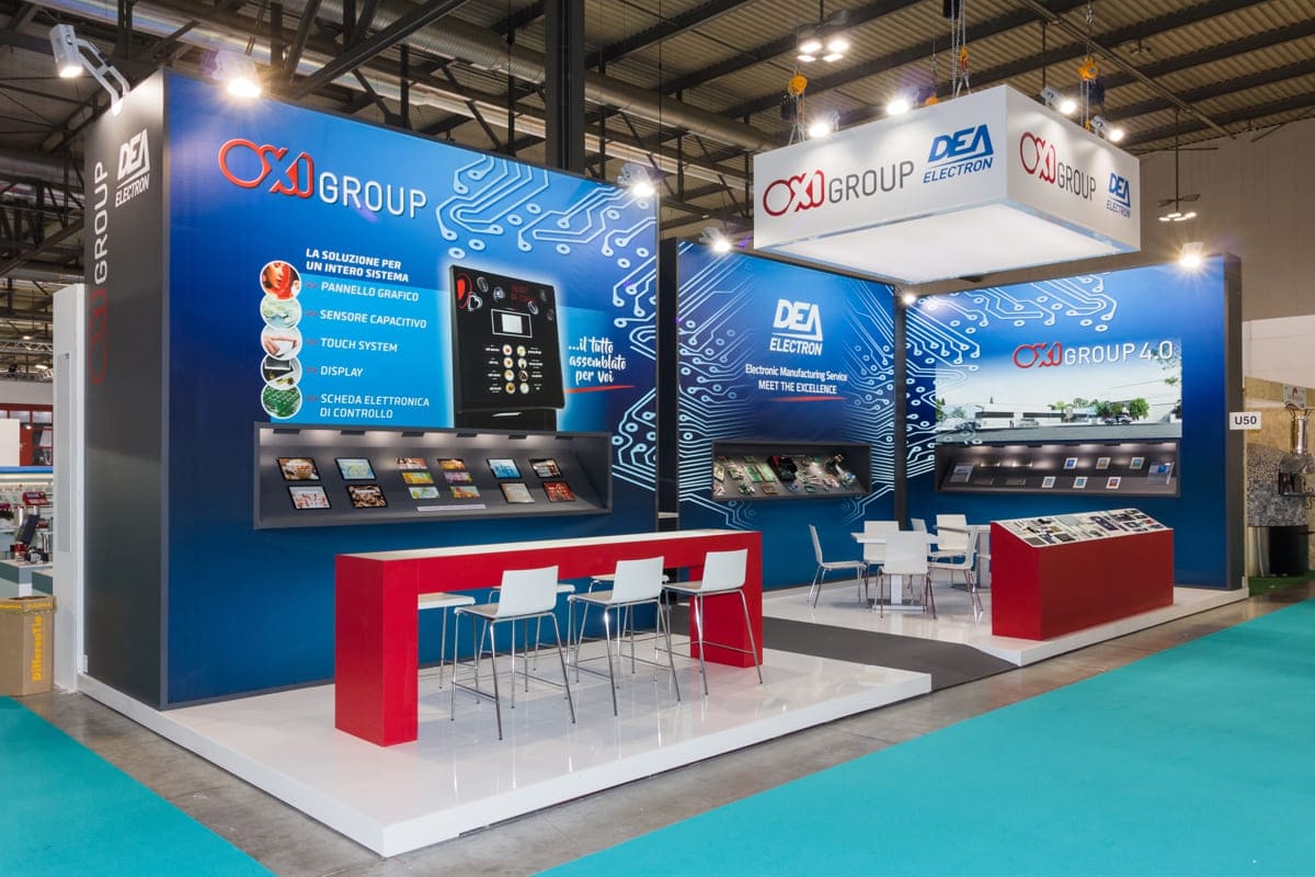 Allestimenti fieristici Host Milano casi reali: l' allestimento stand realizzato per Oxi Group L' immagine raffigura un caso reale di allestimento fieristico: l' allestimento dello stand OXI Group realizzato in occasione di una precedente edizione della fiera Host di Milano. Lo stand è stato allestito su un pavimento rialzato e laccato in colore bianco, con corsia ingresso colore grigio scuro. Per l' allestimento delle parti sono state previste tre grandi box con frontale grafico retroilluminato al centro dei quali sono state ricavate altrettante vetrine espositive per i prodotti. Un grande tavolo alto che funge da zona ricezine ed alcuni tavolini all' interno fromano l' arredamento. Lo stand è completato da un' insegna con loghi, sospesa al centro dell' area.