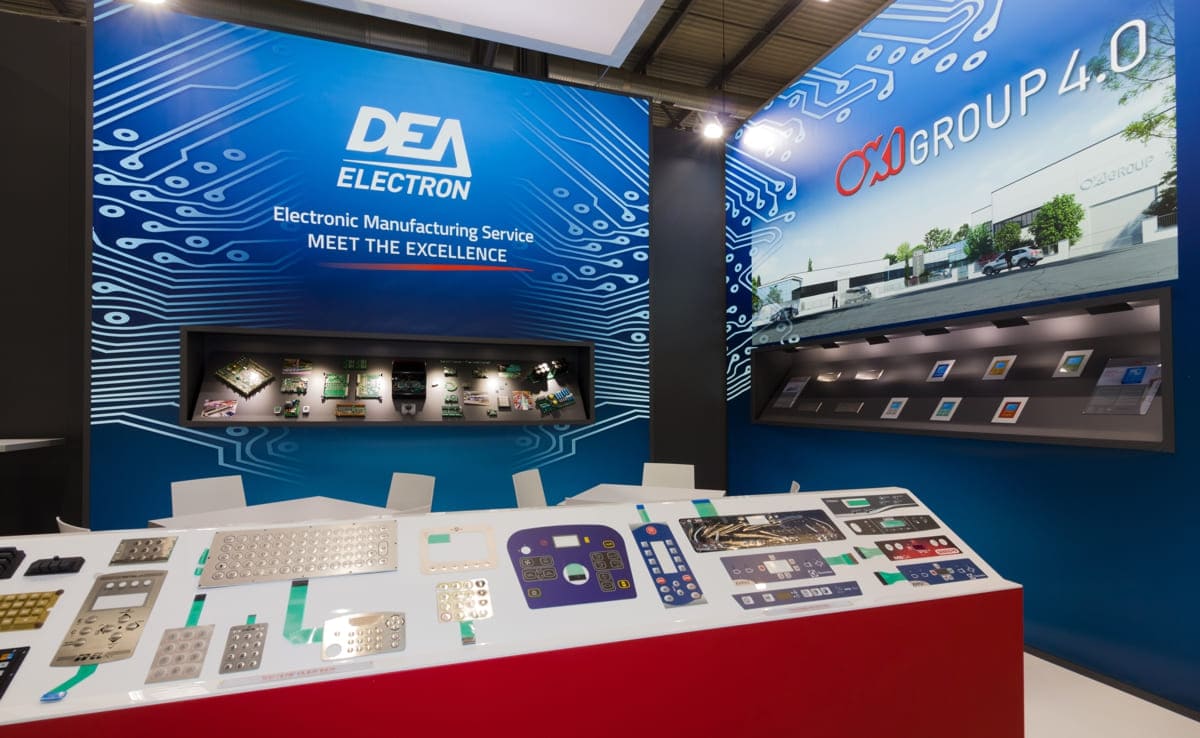 Allestimenti fieristici Host Milano casi reali: l' allestimento stand realizzato per Oxi Group L' immagine raffigura un caso reale di allestimento fieristico: l' allestimento dello stand OXI Group realizzato in occasione di una precedente edizione della fiera Host di Milano. Lo stand è stato allestito su un pavimento rialzato e laccato in colore bianco, con corsia ingresso colore grigio scuro. Per l' allestimento delle parti sono state previste tre grandi box con frontale grafico retroilluminato al centro dei quali sono state ricavate altrettante vetrine espositive per i prodotti. Un grande tavolo alto che funge da zona ricezine ed alcuni tavolini all' interno fromano l' arredamento. Lo stand è completato da un' insegna con loghi, sospesa al centro dell' area.
