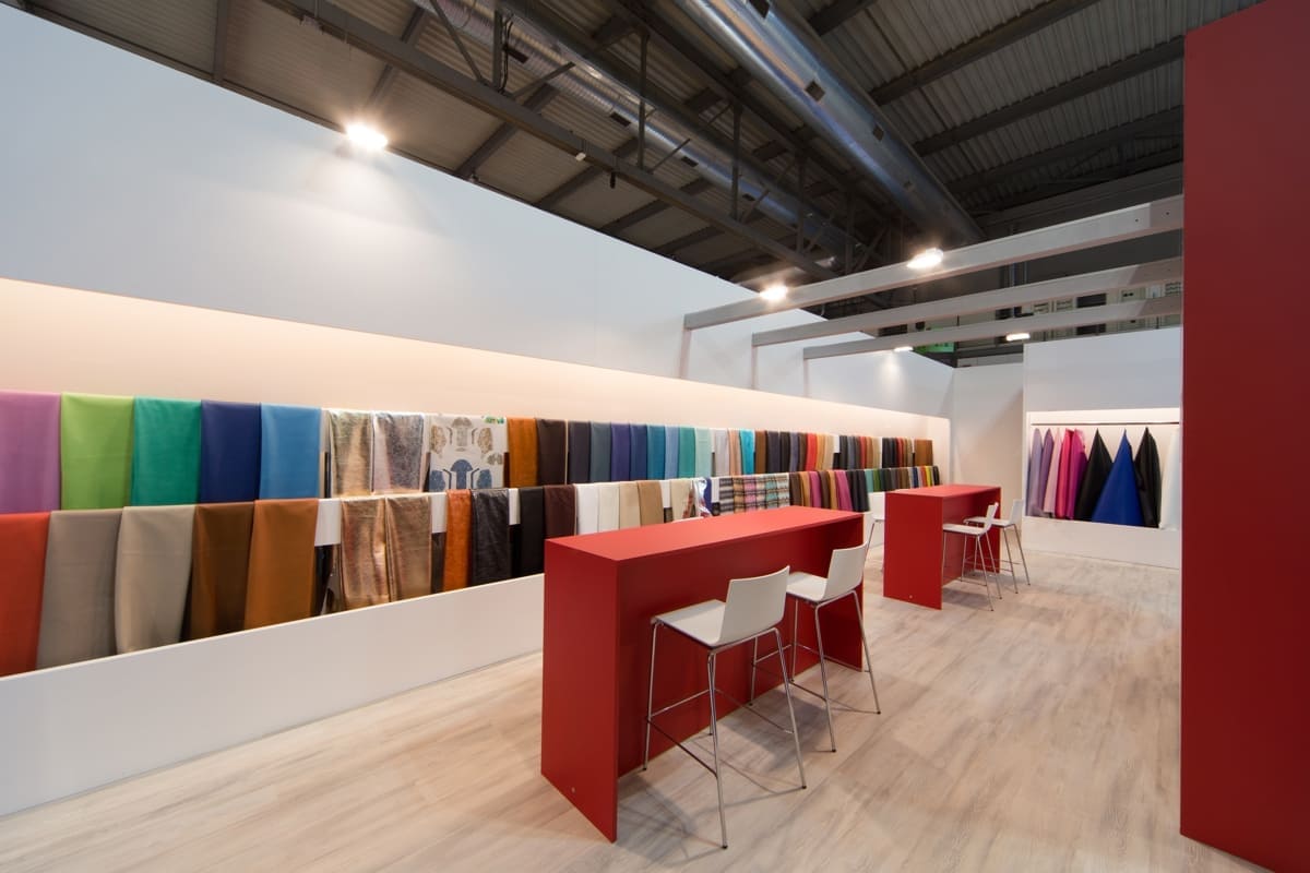 In questa immagine vediamo l' allestimento dello stand Moden Pelli realizzato Lineapelle di Milano fotografato dall'interno con una panoramica sui tavoli squadrati, sull' esposizione a parete e sulle travature al soffitto. Si coglie inoltre molto bene l' effetto cromatico cui dà luogo l' accostamento tra pareti bianche e rosse e il pavimento in essenza nordica rovere grigio.