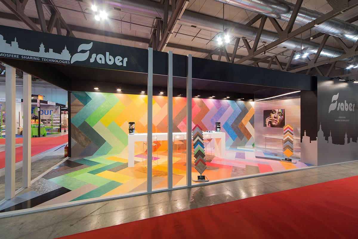 Allestimenti fieristici Madeexpo Milano casi reali: l' allestimento stand realizzato per Saber Color Srl visto da Sud Come la prima, anche in questa foto è raffigurato il caso reale dell' allestimento stand Saber Color realizzato da Diquadro allestitori fiere in occasione di una precedente edizione di Madeexpo di Milano. Lo stand è è qui ripreso dall' esterno. Un punto di osservazione che mostra come lo stand si stato allestito come una scatola che fuori è omogenea e scura e dentro brilla per effetto dei prodotti colorati esposti.