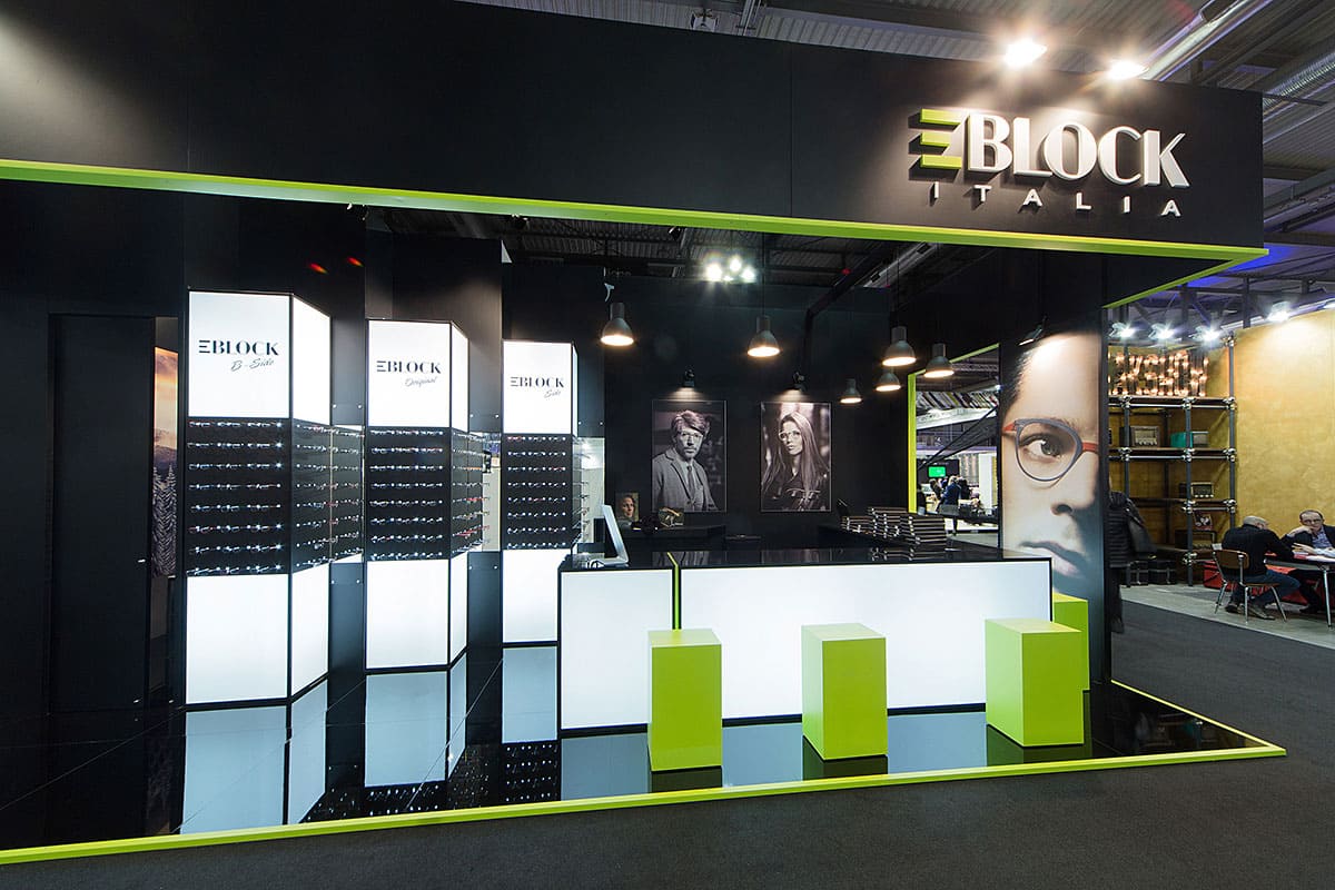Allestimenti fieristici Mido Milano casi reali: l' allestimento stand realizzato per E-Block di Som Occhiali Srl visto da Sud La terza foto dell' allestimento dello stand eblock realizzato da Diquadro per la fiera Mido di Milano mostra lo stand ripreso frontalmente. In questa foto dello stand si coglie bene la forma e lo stile del punto di accoglienza e delle relative sedute che si rifanno alle forme squadrate del logo e-block