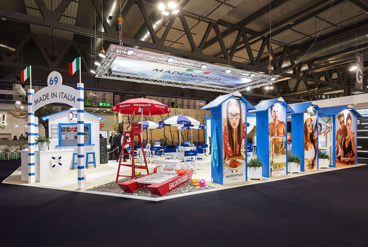 L' immagine raffigura un caso reale di allestimento fieristico: l' allestimento dello stand Made in Italia dell'azienda L'amy Group realizzato da Diquadro in occasione di una precedente edizione della fiera Mido di Milano. L' immagine ci mostra una panoramica esterna dello stand presa dal lato sinistro, cio Sud Ovest. È ben visibile la zona allestita con sabbia marina, moscone, sedia bagnino e cabine, nonché ingresso e cabina bar.