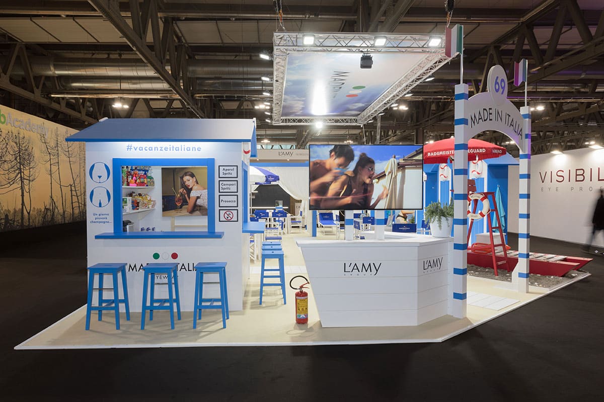In quest'altra foto vediamo una panoramica sul lato Ovest dell' allestimento fieristico realizzato per Made in Italia by L'amy Group al Mido di Milano. L'inquadratura mostra in particolare la cabina uso bar con gli sgabelli e poi l'ingresso alla sua destra.
