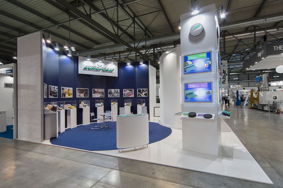 L'immagine raffigura un caso reale di allestimento fieristico: l'allestimento dello stand Eurofoam Technical Rubber realizzato in occasione di una precedente edizione della fiera Plast di Milano. La foto mostra una vista panoramica presa da Ovest nella quale si vede con chiarezza una delle due pareti curve e il totem con l'esposizione congiunte per entrambe le aziende.