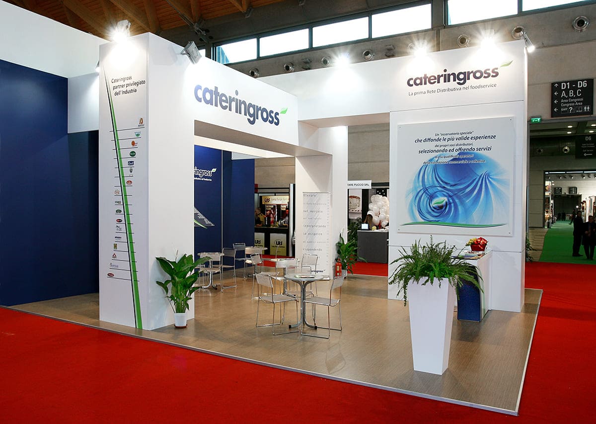 L'immagine raffigura un caso reale di allestimento fieristico: l'allestimento dello stand Cateringross realizzato per fiera Sigep di Rimini. L'immagine ci mostra una vista da Ovest dello stand realizzato con panoramica sul banco ricezione, sulla zona tavoli e sulle parti grafiche.