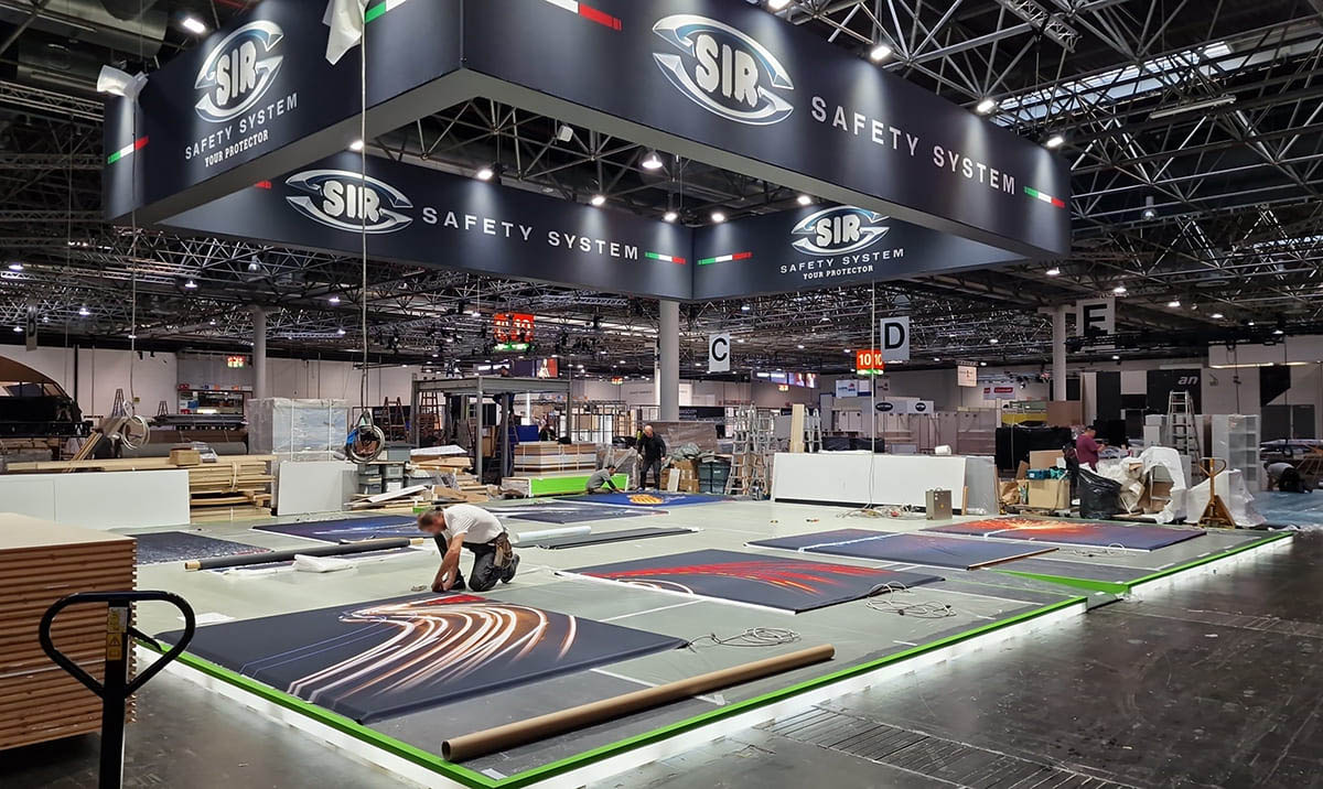 Allestimenti stand per fiera A+A Dusseldorf sulla sicurezza · Un esempio di allestimento stand fieristico in fase di montaggio in cantiere L'immagine mostra una fase del montaggio in cantiere di uno stand realizzato per la fiera A+A di Dusseldorf, dedicata alla sicurezza sul lavoro. Nella foto si vede il personale mentre è intento a posare teli grafici in sicurezza