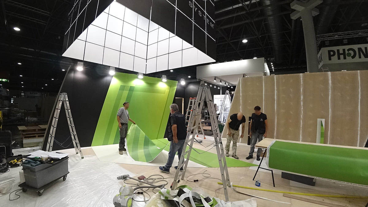 L'immagine mostra una fase dell'allestimento di uno stand per fiere personalizzato con wrapping. Nella foto si vede il personale al lavoro mentre è intento a wrappare le suprefici dello stand con teli grafici adesivi