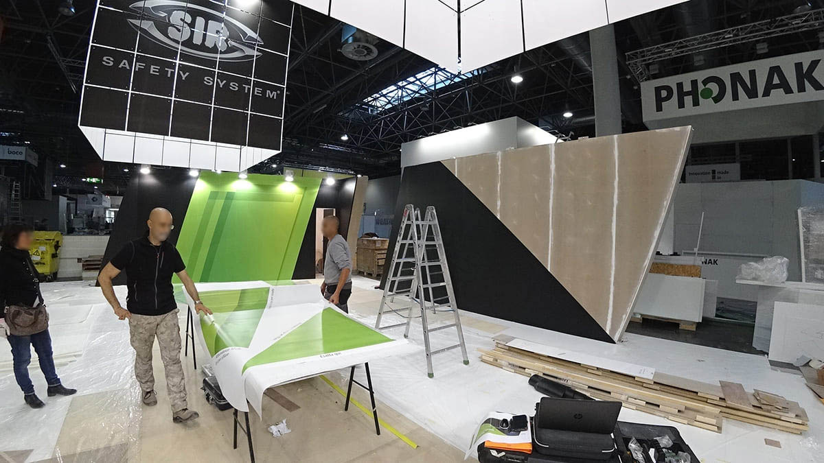 L'immagine mostra una fase di montaggio e decorazione di uno stand fieristico personalizzato con wrapping. Nella foto si vede il personale al lavoro mentre è intento a preparare il materiale con cui wrappare le suprefici dello stand