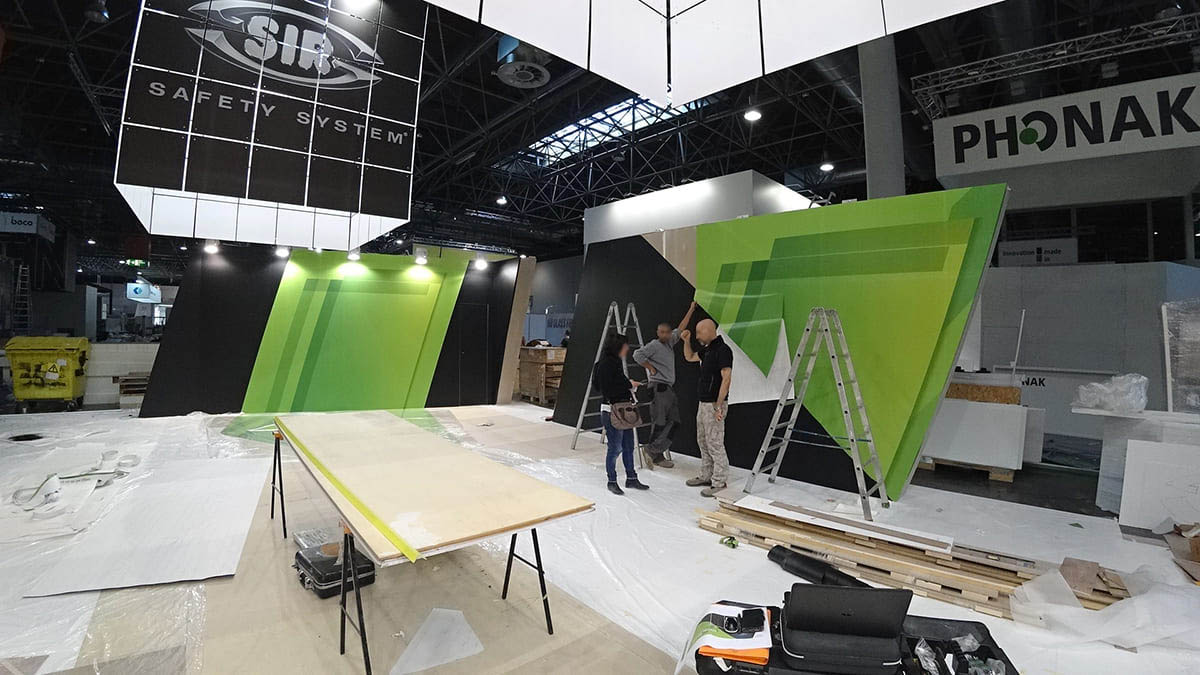 L'immagine mostra un'altra fase dell'allestimento di uno stand per fiere personalizzato con wrapping. Nella foto si vede il personale al lavoro mentre è intento a confrontarsi sulle oprerazioni di wrappatura delle suprefici