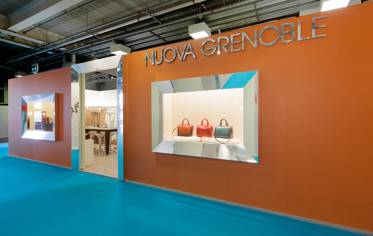 Allestimenti stand per fiere · Un esempio di allestimento stand per fiera L'immagine mostra l'esterno di un allestimento stand per fiera: la zona dell'ingresso. Nella foto di vede la parete frontale esterna dello stand caratterizzata dalla presenza di due ampie vetrine espositive ai lati dell'ingresso che si trova al centro. Sia l'ingresso che le due vetrine sono rese particolari dall'allestimento di tre grandi cornici in acciaio semilucido e e di un logo aziendale parimenti realizzato in acciaio.