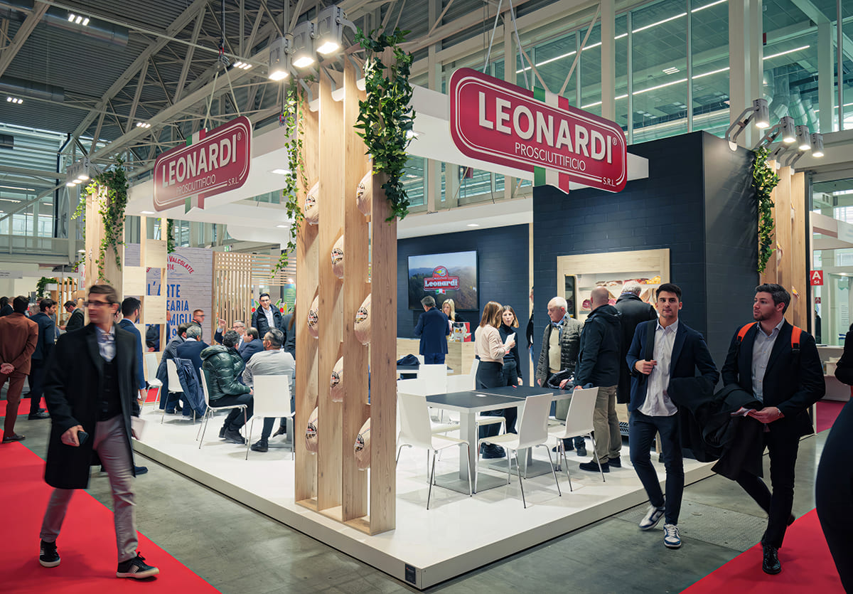 Allestimenti stand per fiere · Un esempio di allestimento stand per fiera L'immagine mostra un particolare di un allestimento stand per fiera durante il suo svolgimento: la zona esterna. Nella foto di vedono, oltre alle persone in visita allo stand, in primo piano due grandi espositori per gli articoli prodotti dall'azienda. Poi, sul fondo, la zona riservata con ripostiglio, uffici, tavoli per gli incontri informali, il banco ricezione.