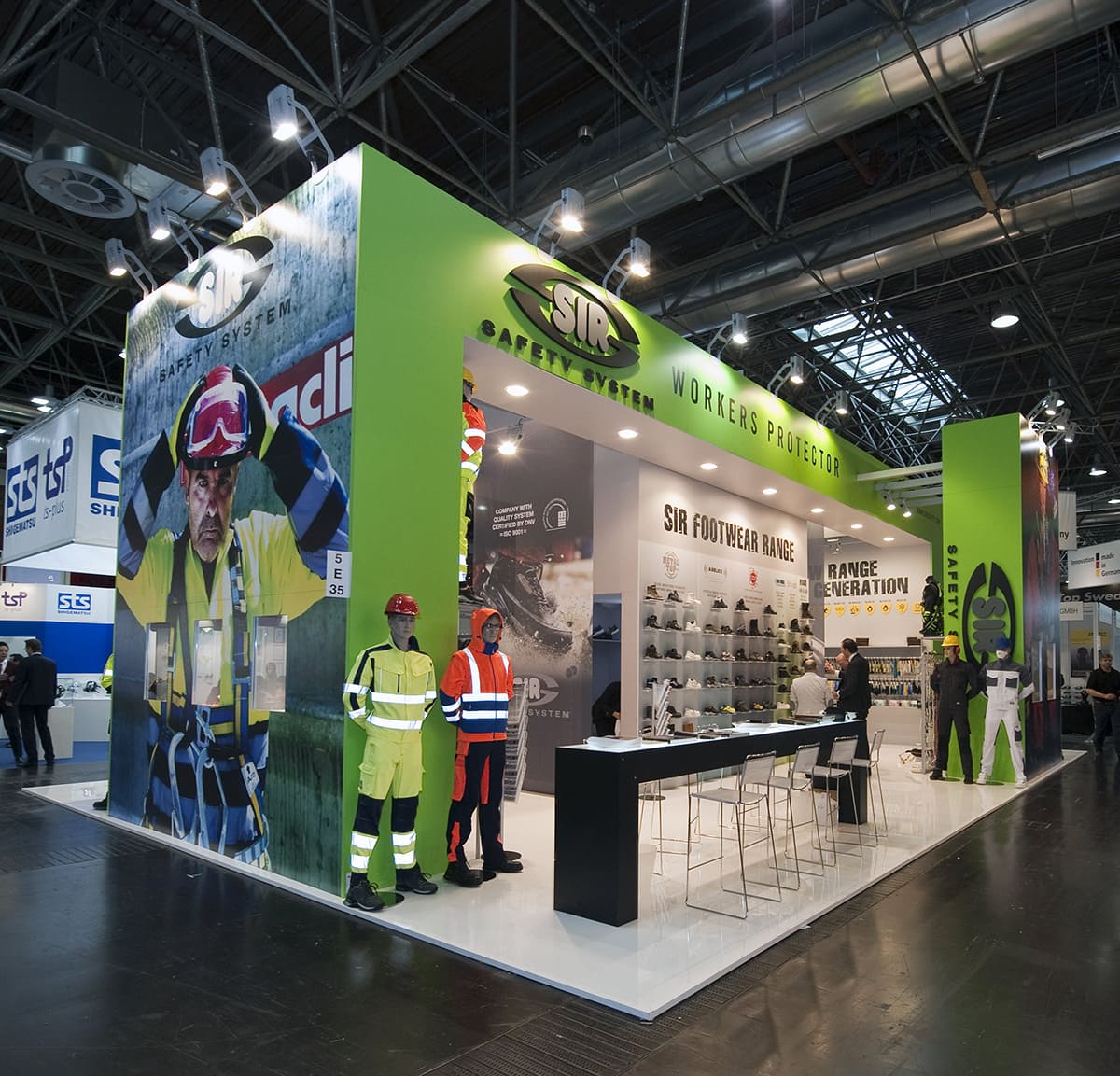 L'immagine mostra un allestimento realizzato da Diquadro per la fiera A+A di Düsseldorf, allestito su un pavimento laccato bianco la cui struttura si articola anzitutto in un imponente ponte centrale a due travi che poggiano ai lati su due volumi strutturali, all'interno dei quali sono state realizzate vetrine espositive. Poi si articola in un box centrale che caratterizza lo spazio espositivo e regge, al centro, le due grandi travi. Infine si articola in altri due volumi ad isola che si collocano agli angoli opposti dell'area. Completano l' allestimento stand alcune light box disposte a pettine sul lato Nord ed un grande tavolo alto retroilluminato sul lato sud.