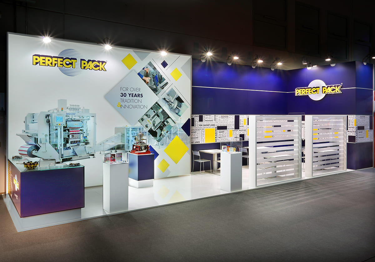 Allestimenti stand fiera Achema Francoforte · Un esempio di allestimento stand fieristico realizzato: guarda come l'abbiamo sviluppato L' immagine mostra un'allestimento realizzato da Diquadro per la fiera Achema di Francoforte. Lo stand raffigurato è allestito su un pavimento rialzato e laccato in colore bianco, sul quale sono erette le paredi fondo caratterizzate da una vivace lavorazione grafica sui toni di bianco e giallo su campo blu. Sul lato destro è presente un generoso box dedicato allo stivaggio delle scorte; a fianco a questo, una zona di incontri con tavolini, schermata da una coppia di diaframmi semitrasparenti. La zona di accoglienza si trova sulla sinistra ed è caratterizzata dalla pesenza di un banco ricezione alle cui spalle è ricreata una grande superficie grafica retroilluminata con motivi che riprendono quelli della parete di fondo.