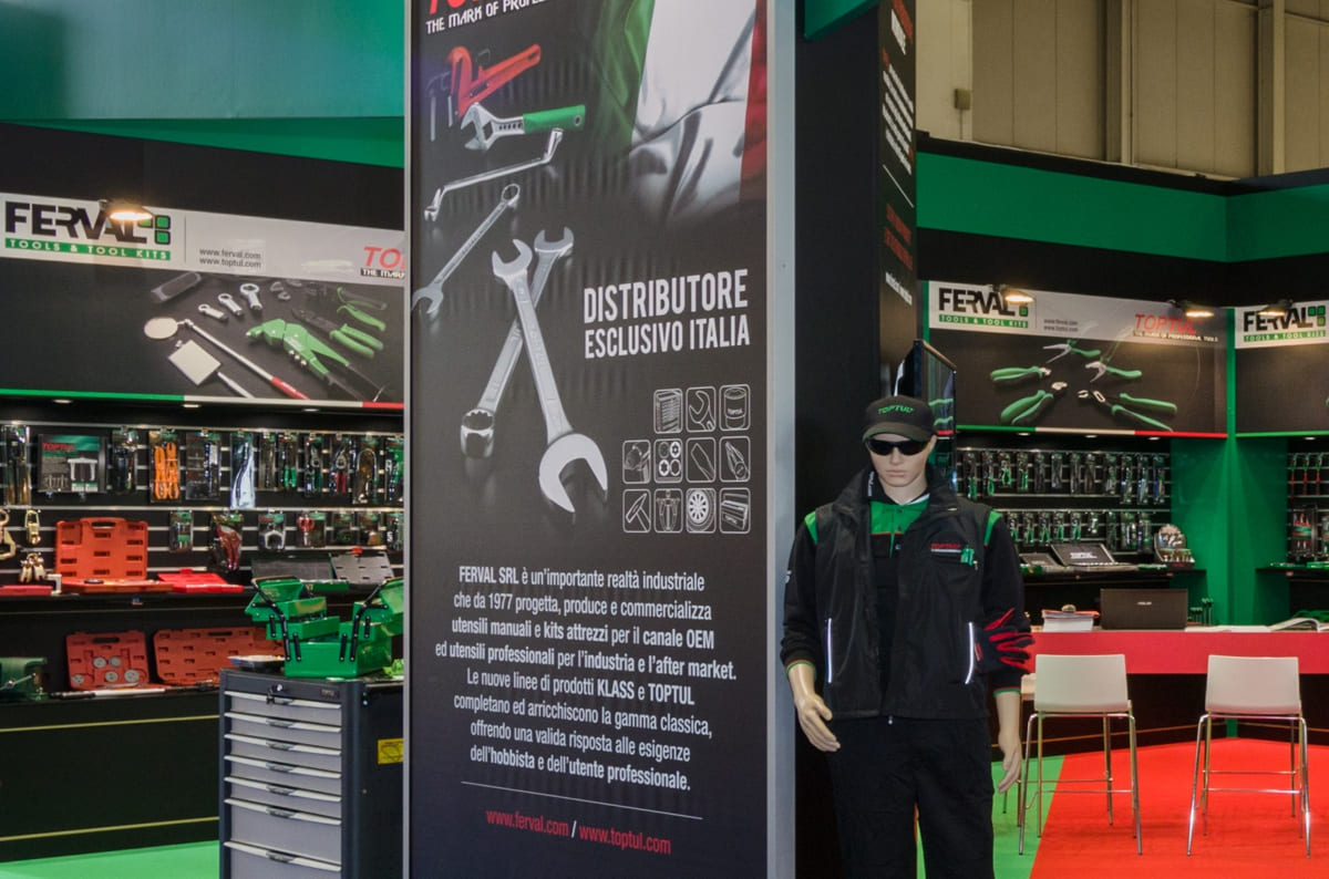 Allestimenti stand fiera Autopromotec Bologna · Un esempio di allestimento stand fieristico realizzato: guarda come l'abbiamo sviluppato L'immagine mostra un allestimento realizzato da Diquadro per la fiera Autopromotec di Bologna. Si tratta dello stand di un'azienda che partecipa a questa fiera dove espone attrezzature per lavori di meccanica e kit di pronto intervento meccanico. La foto ci mostra una panoramica dell'allestimento presa dall'esterno, a destra. Si nota sui due lati di fondo lo sviluppo delle pareti alte quattro metri alla base delle quali è stata ricreata un'ampia zona espositiva per i singoli utensili e per i vari kit. Particolare evidenzia la foto la riserva poi alla struttura di allestimento esterno dello stand, caratterizzata dalla particolare forma delle travature frontali che si rifanno alle forme delle travi in ferro degli stabilimenti industriali dei quali si è voluto qui richiamare l'idea.
