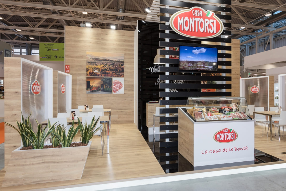 L'immagine mostra un esempio di allestimento realizzato da Diquadro per la fiera Cibus di Parma. Si tratta di uno stand per fiere allestito su un pavimento rialzato con zoccolo retroilluminato, rivestito in rovere naturale. La struttura è formata da una parete di fondo rivestita anch'essa in rovere naturale e con un box magazzino centrale rifinito in bainco e nero lucido. La zona centrale è caratterizzata dalla presenza di una struttura a ponte lavorata a doghe di colore bianco e nero. Ai lati di questa struttura si trovano le due zone trattative con tavoli che sono schermati da diaframmi semitrasparenti. Completano l'allestimento di questo stand un'esposizione di prodotti in vetrina refrigerata fdrontale ed un paio di separé con piante verdi.