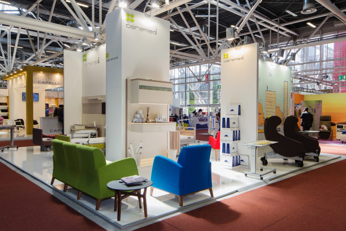 Allestimenti stand fiera Exposanità Bologna · Un esempio di allestimento stand fieristico realizzato: guardate come lo abbiamo sviluppato L'immagine mostra un'allestimento realizzato da Diquadro per la fiera Exposanità di Bologna. Si tratta dello stand di un'azienda che partecipa ad Exposanita dove espone ausili medici per l'assistenza agli anziani, alle persone bisognose di assistenza medica e, in generale, alle disabilità. Lo stand raffigurato è allestito su un pavimento rialzato di colore bianco. La struttura verticale riproduce fedelemente gli spazi e le pareti di una camera di degenza, arredata con i prodotti dell'azienda committente. Particolare attenzione la foto la pone sulla rifinitura delle pareti dello stand, allestite per mezzo del wrapping di tessuti murali decorati.