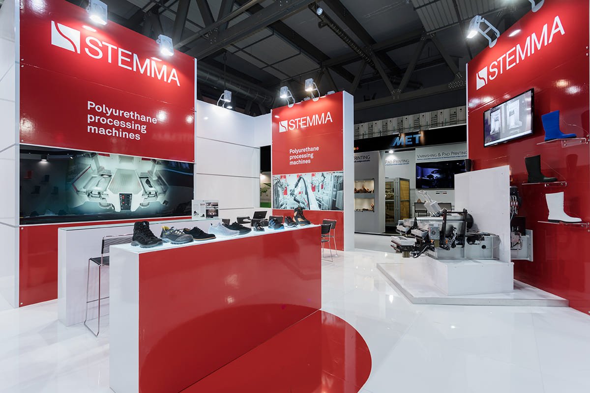 Allestimenti stand per fiera Simac Tanning Tech di Milano · Un esempio di allestimento stand fieristico realizzato: guarda come l'abbiamo sviluppato L'immagine mostra un allestimento realizzato per fiera Simac Tanning Tech Milano. L'immagine raffigura una vista di tre quarti dello stand allestito per accogliere i clienti ed esporre alcune macchine per lo stampaggi odi suole e calzature. Per allestire lo stand lo spazio è stato delimitato con quattro grandi colonne su cui campeggiano scritte, foto e da altri elementi grafici che spiegano la produzione di macchine per la lavorazione di calzature e pelletteria. La zona interna centrale dell' allestimento fieristico è in parte dedicata alla zona incontri e in parte dedicata all'esposizione dei macchinari.