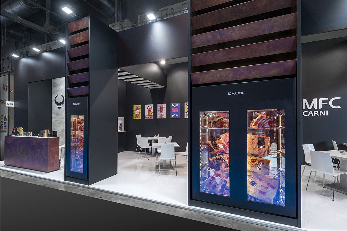 Allestimenti stand per fiera Tuttofood Milano · Un esempio di allestimento stand fieristico realizzato: guarda come l'abbiamo sviluppato L'immagine mostra un'allestimento realizzato da Diquadro per la fiera Tutto Food di Milano. Questo stand fieristico è allestito su un pavimento rialzato e laccato in colore bianco. La struttura di allestimento è realizzata con pareti alte quattro metri di colore nero. Sono ricreati una cucina sulla sinistra ed uno spazio per gli incontri con tavolini sulla restante parte dell'area. Il fronte dello stand è costituito da tre grandi box dentro i quali sono incassati degli eleganti frigoriferi per l'esposizione dei prodotti alimentari. Questi box sono collegati strutturalmente con la parete di fondo per mezzo di cassonetti di colore nero, rifiniti inferiormente con doghe in colore cemento. Un grande banco ricezione a sinistra è rifinito in colore ruggine e si staglia su una parete in cemento. Lo stand è completato da grafica e loghi.